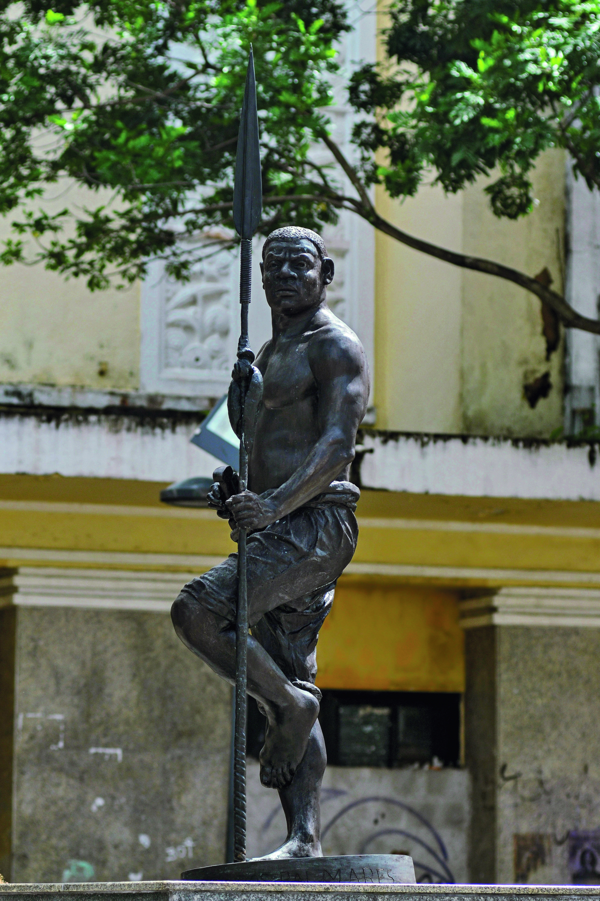 Fotografia. Uma estátua de bronze de um homem de cabelo curto, sem camiseta, com calça rasgada e segurando uma lança.