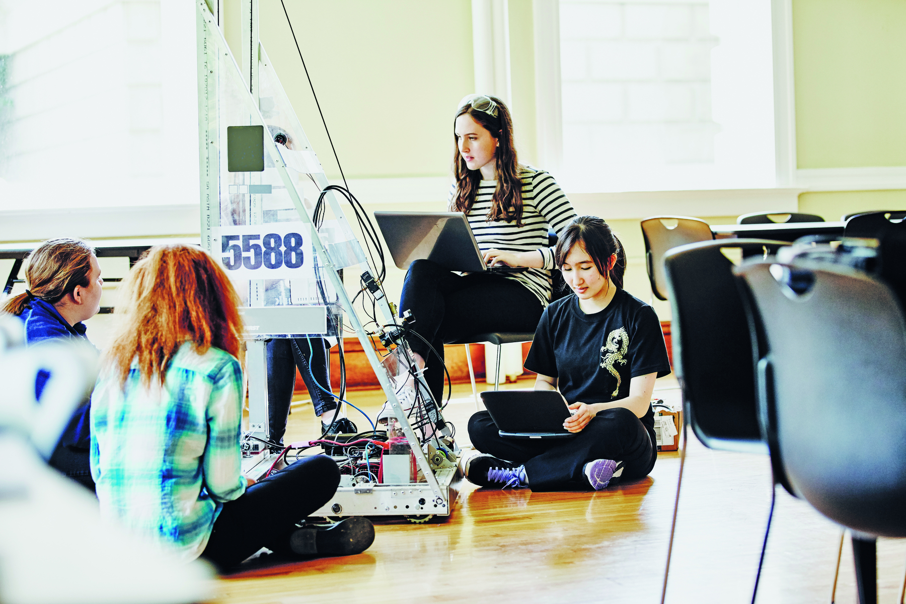 Fotografia. Quatro jovens sentadas em uma sala. Elas estão com laptops no colo. À frente delas há um robô.