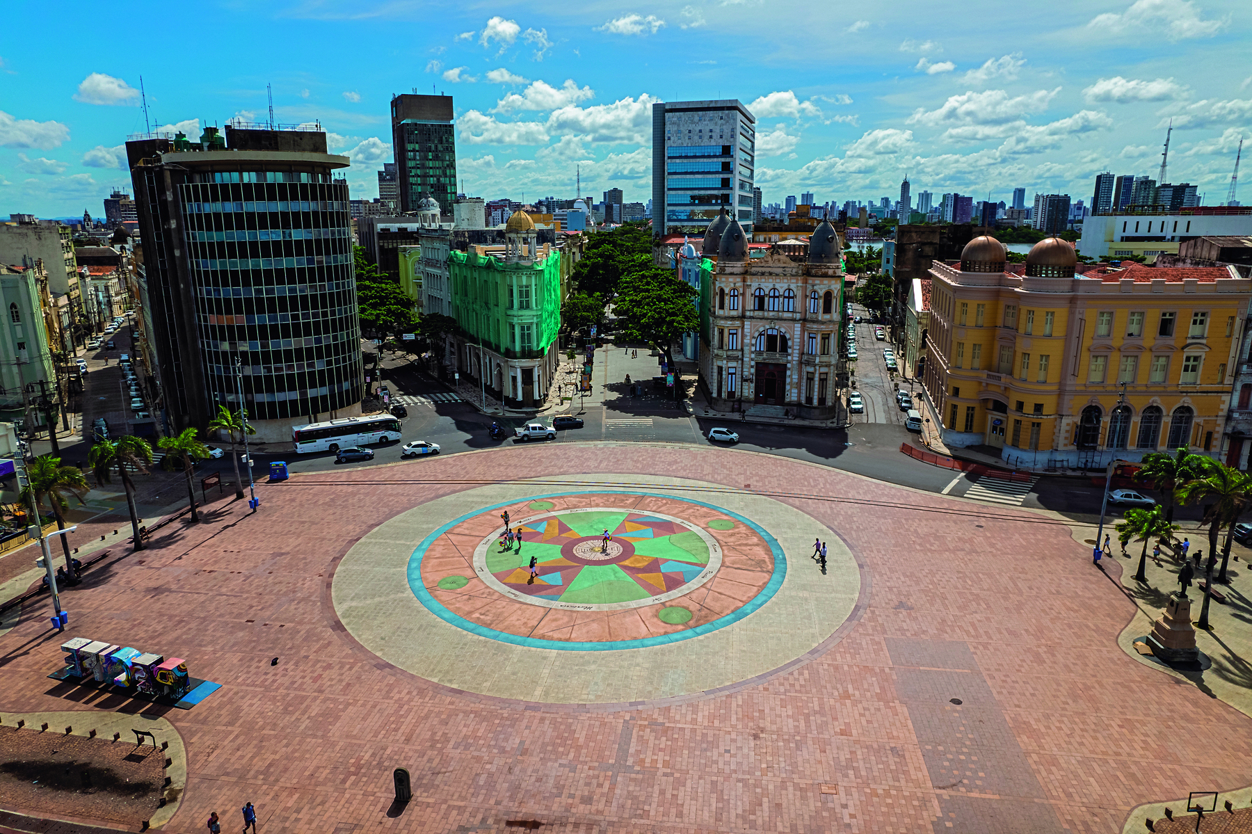 Fotografia. Um espaço público largo com um círculo no centro. Ao redor há prédios e ruas.