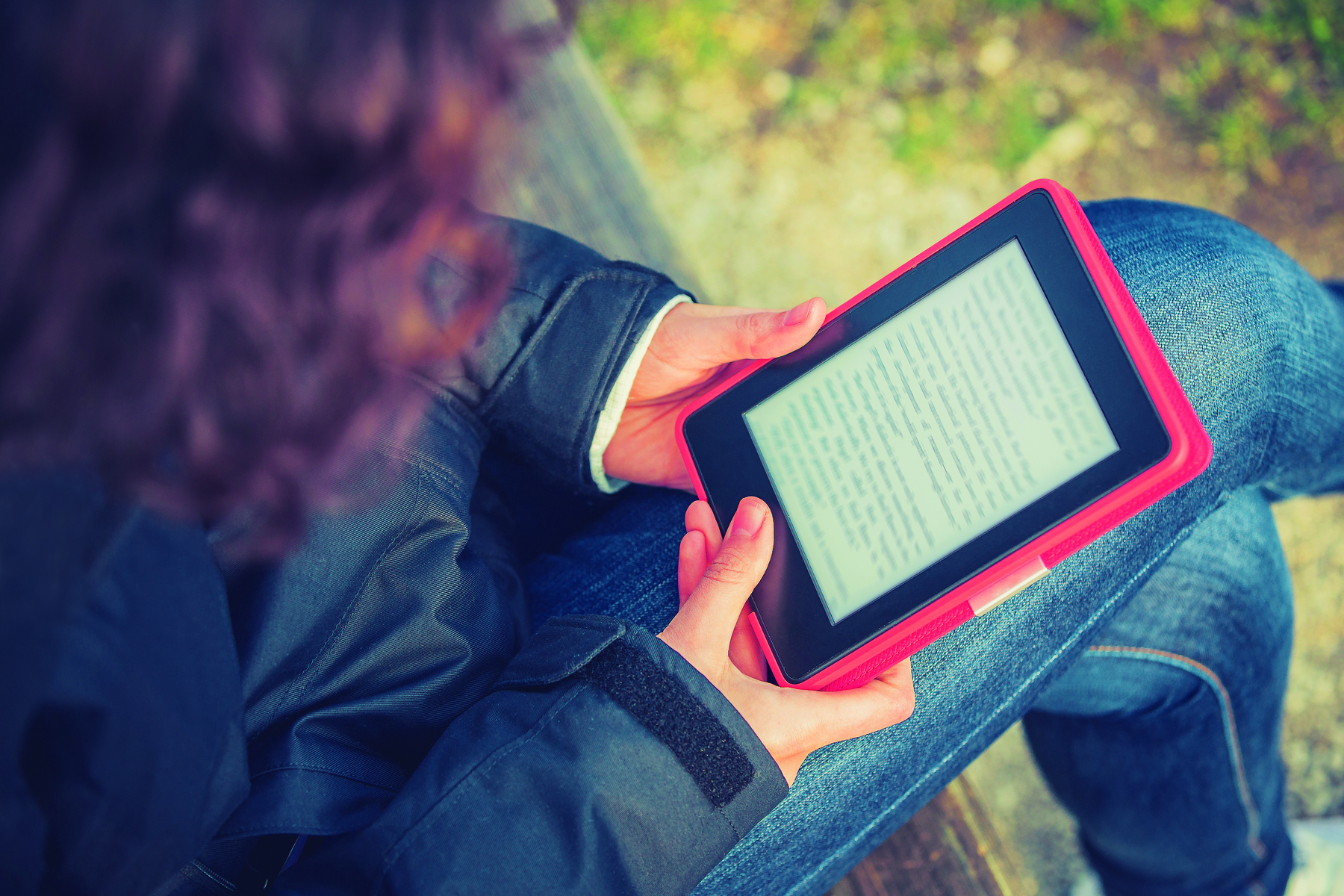 Fotografia. Uma menina usando calças jeans e casaco preto segurando com as duas mãos um leitor de livros digital preto com capa vermelha. Na tela do leitor há um texto.