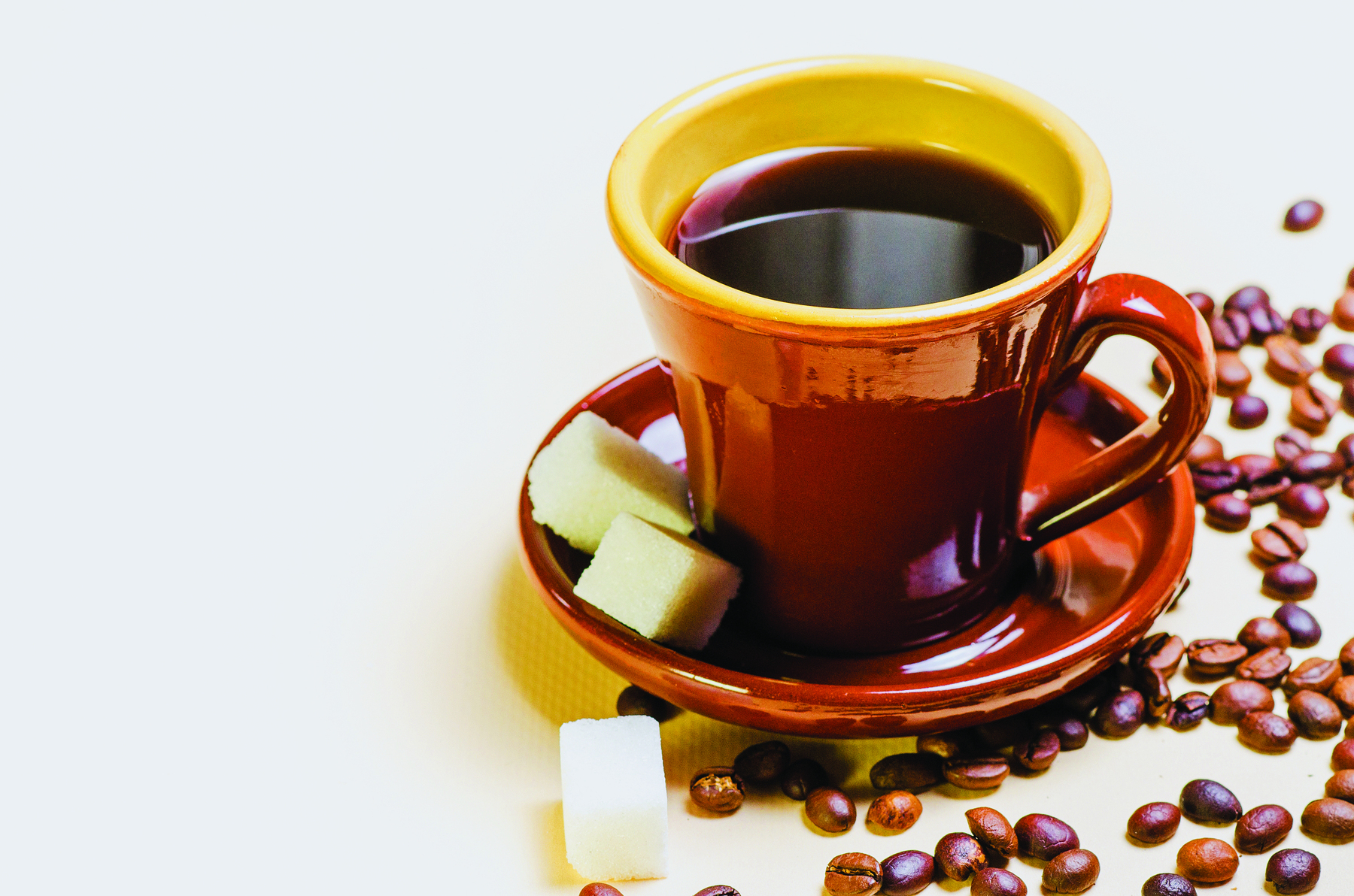 Fotografia. Uma xícara marrom com café sobre um pires marrom. Ao lado, sobre o pires, há dois cubos de açúcar. Ao redor da xícara e do pires, sobre a mesa, há grãos de café e mais um cubo de açúcar.