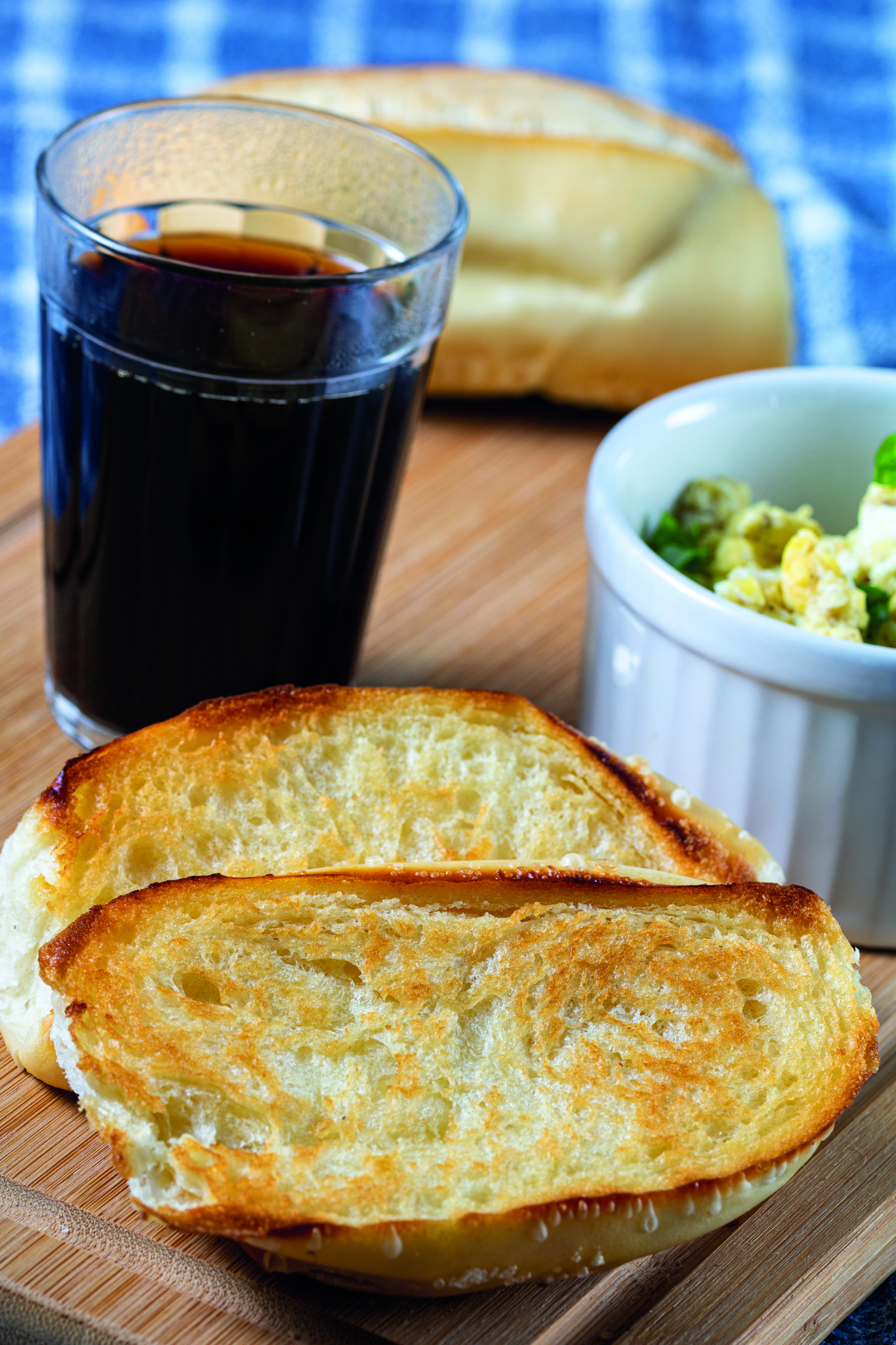 Fotografia. Duas fatias de pão francês torrado e um copo de vidro com café. Ao lado há uma tigela com ovo mexido.