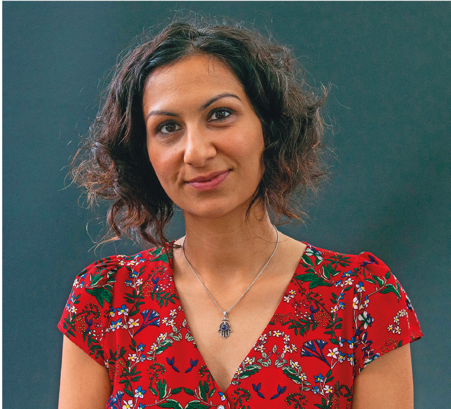 Fotografia. Yasmin Khan, uma mulher de cabelo castanho, cacheado e curto, sobrancelhas finas, usando colar e uma blusa vermelha com estampa verde e branca.