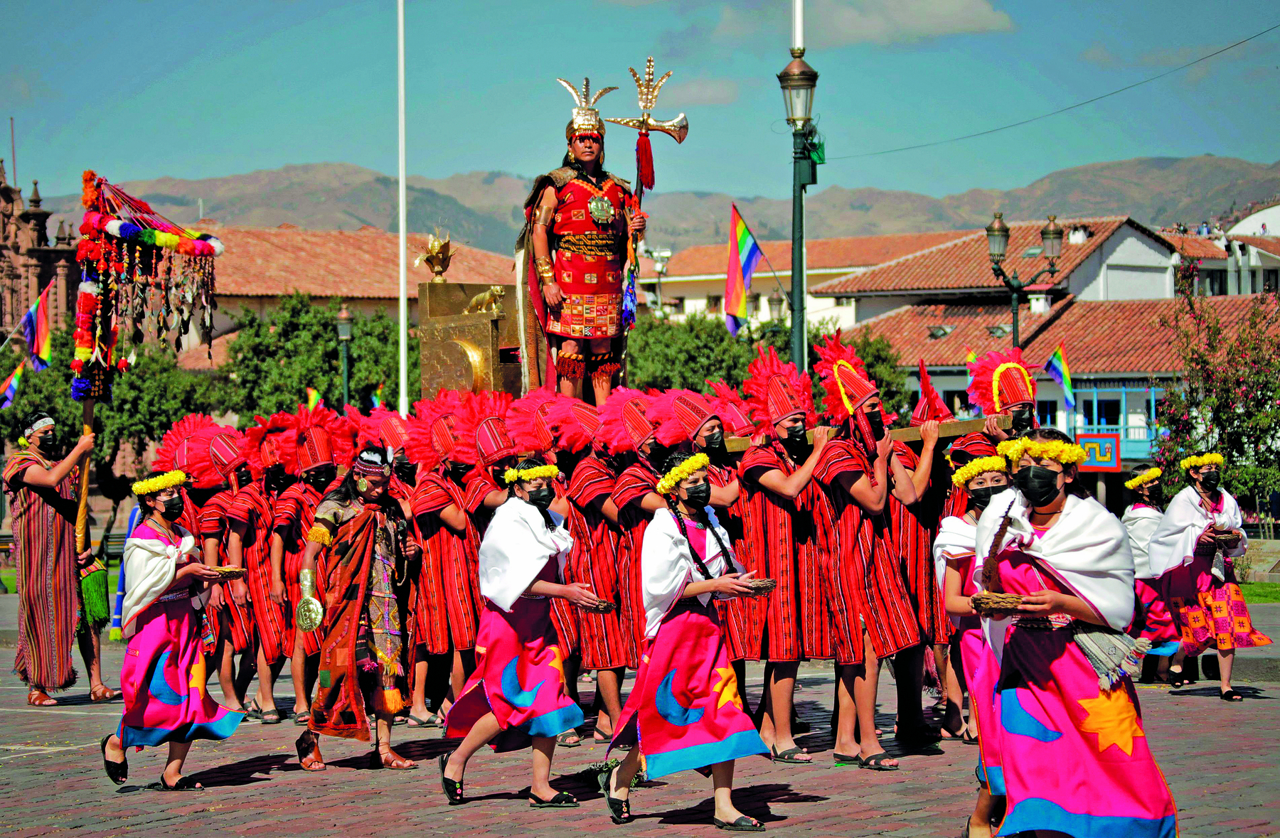 Fotografia. Diversas pessoas usando roupas vermelhas típicas de Inti Raymi com chapéus vermelhos. Algumas delas têm uma coroa de flores amarela e um tecido branco sobre os ombros. Acima delas há um homem com roupas típicas vermelhas, com detalhes dourados, uma coroa dourada e um objeto dourado na mão esquerda.