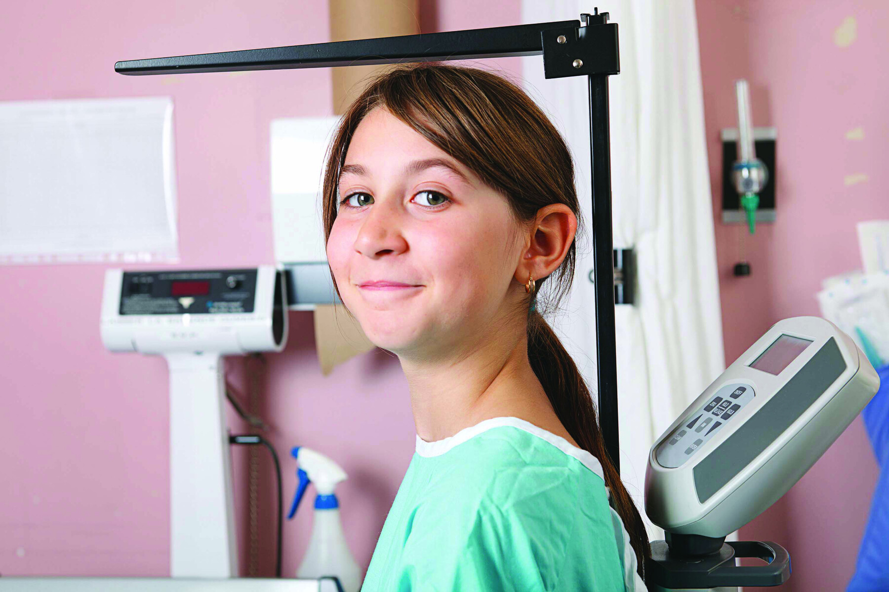 Fotografia. Uma menina adolescente em um consultório médico. Ela está usando um avental hospitalar. Ela está em pé sobre uma balança que tem uma haste para medir a altura. Ao fundo há outros maquinários médicos.