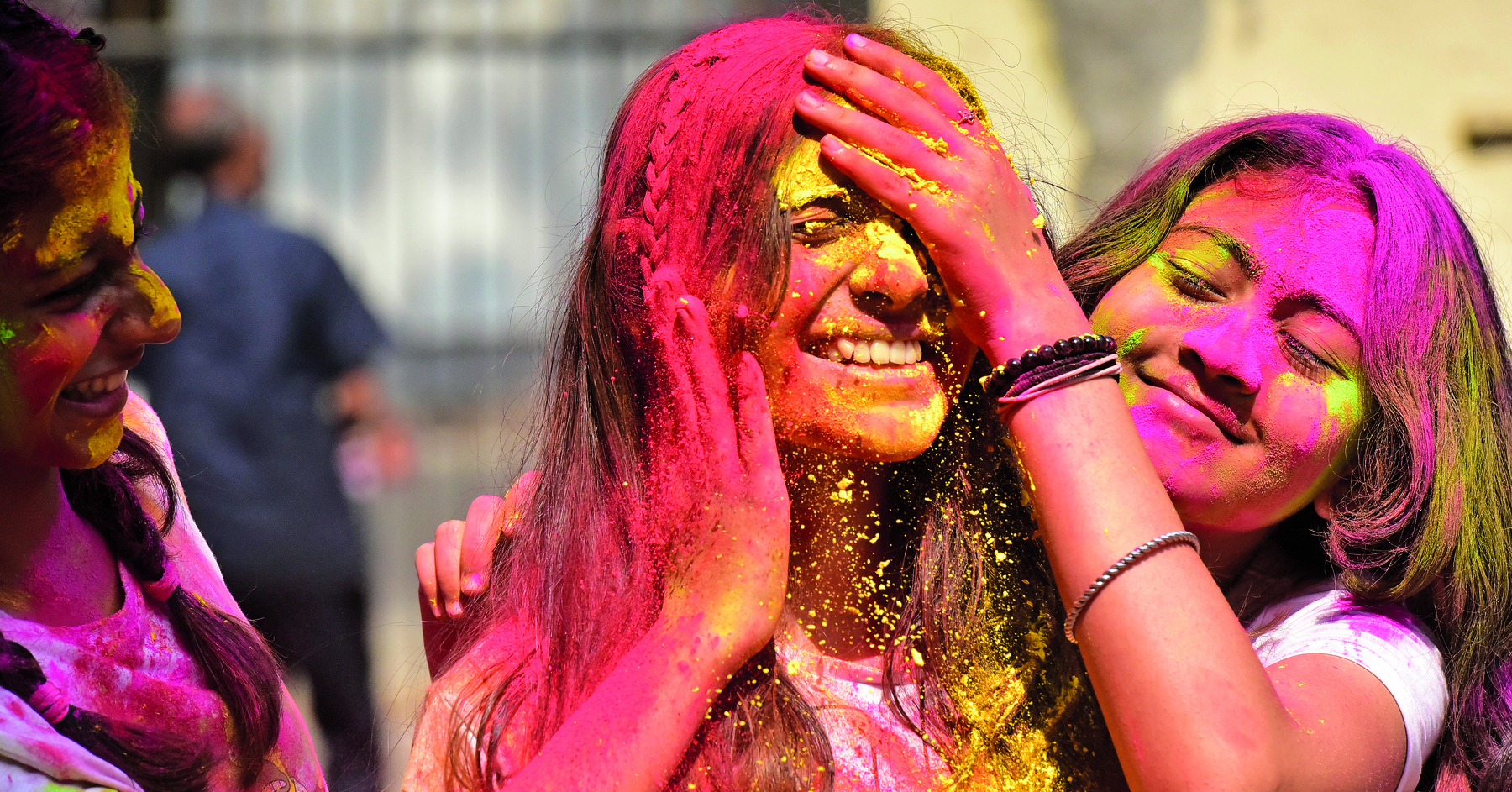 Fotografia. Duas jovens com o cabelo e a pele cobertos com pós coloridos vermelho, amarelo e cor de rosa. Uma está com a mão na testa da outra. As duas estão sorrindo.