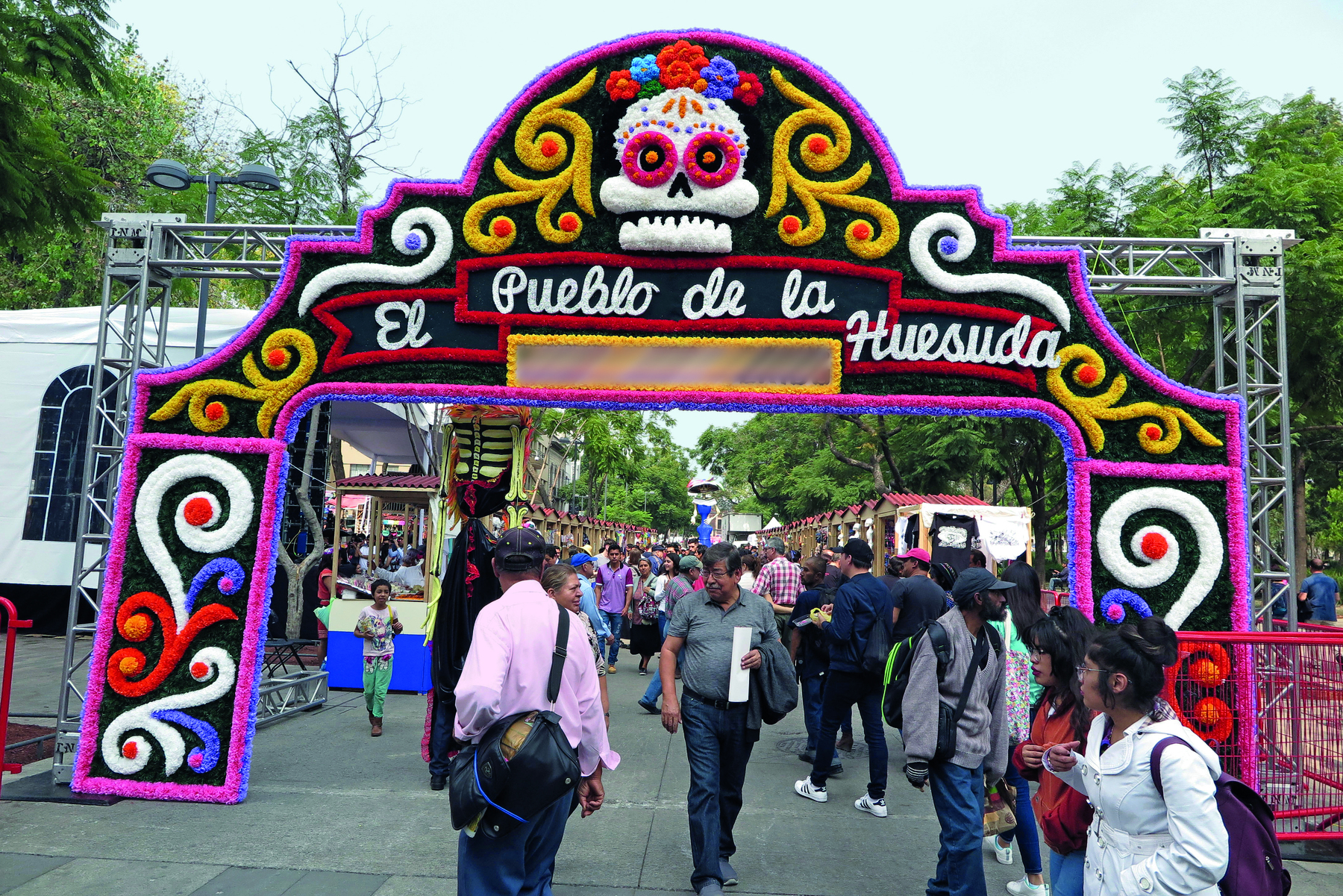 Fotografia. Ao centro há um portal em forma de arco decorado com arabescos coloridos e uma caveira branca, com os olhos vermelhos e flores sobre a cabeça. Abaixo da caveira lê-se El Pueblo de la Huesuda. Abaixo do portal há pessoas caminhando.