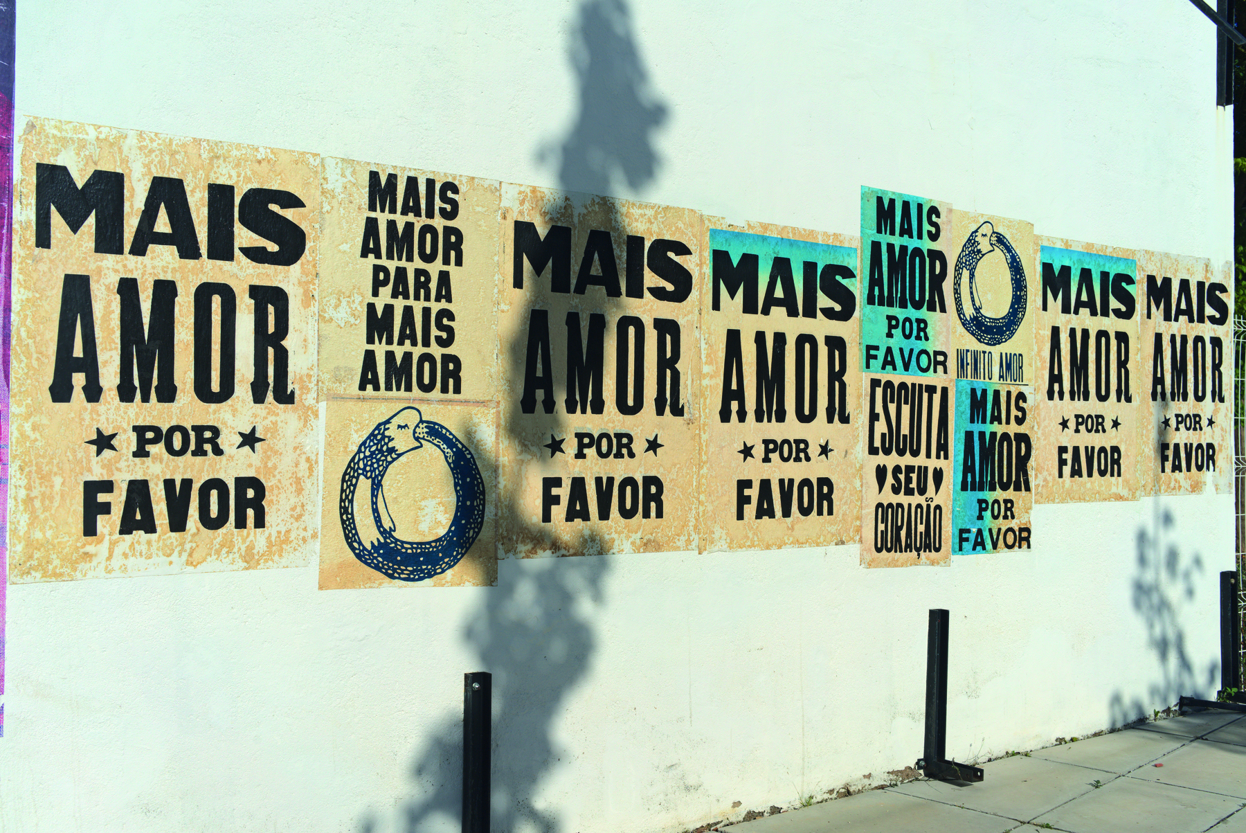 Fotografia. Cartazes colados em um muro. Os cartazes têm um fundo bege e contêm textos em cor preta. Neles, lê-se MAIS AMOR POR FAVOR e ESCUTA SEU CORAÇÃO.