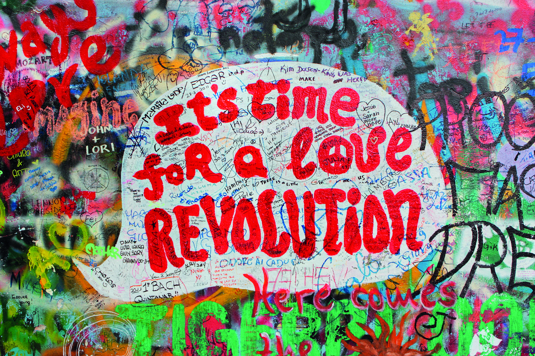 Fotografia. Detalhe do muro decorado com pinturas coloridas. No muro, lê-se IT'S TIME FOR A LOVE REVOLUTION escrito em tons de vermelho. A letra O da palavra LOVE tem o símbolo de paz e amor.