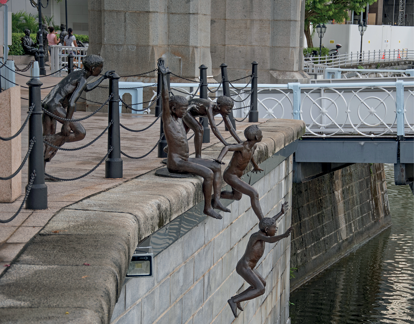 Fotografia. Escultura de bronze de diversas crianças em pé e sentadas à beira de um rio. Algumas estão saltando para dentro do rio.