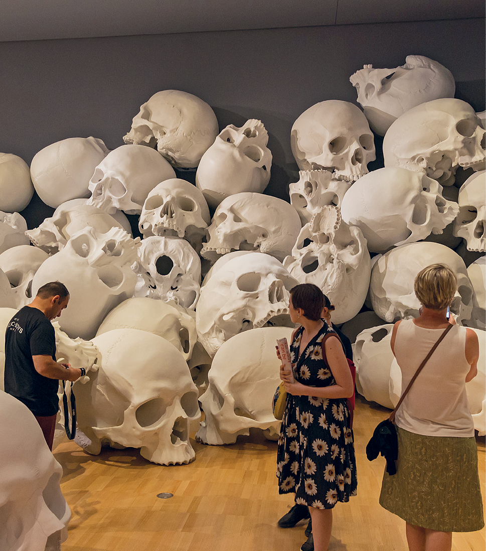 Fotografia. Três pessoas olhando crânios humanos brancos gigantes empilhados uns sobre os outros no chão.