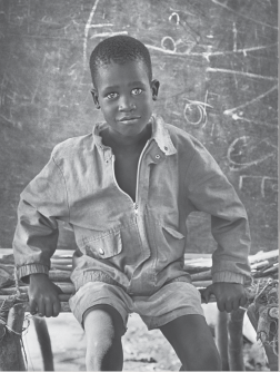 Fotografia. Preto e branco. Um menino negro de olhos claros e cabelo curto vestindo shorts e camisa de manga comprida lisos está sentada em um banco formado por varas cilíndricas. Ela apoia as duas mãos nessa estrutura e olha para a câmera. No fundo há uma parede com riscos brancos.