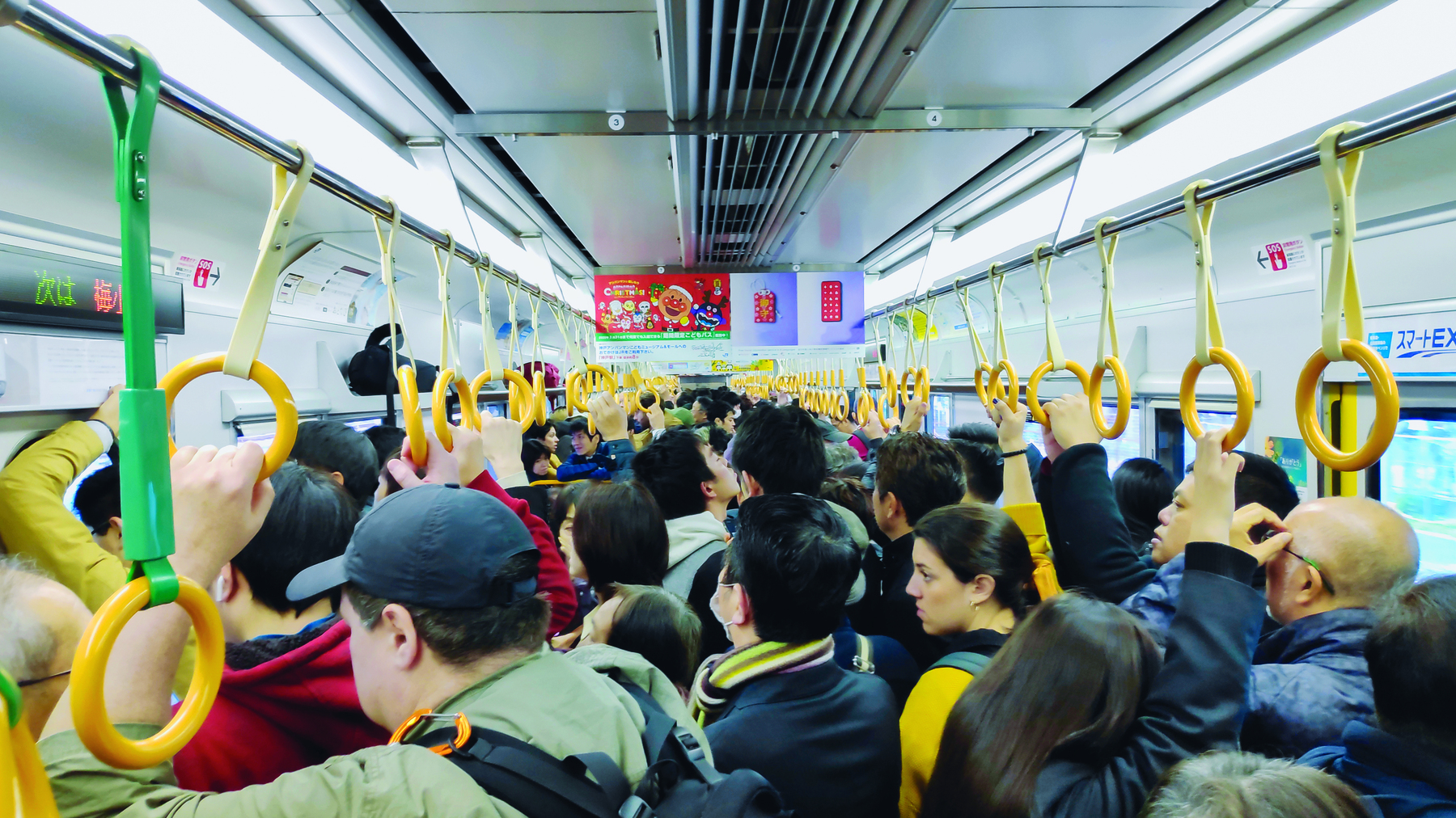 Fotografia. Vagão de metro lotado, com as pessoas segurando argolas amarelas fixadas em uma barra transversal que está próximo do teto do vagão.