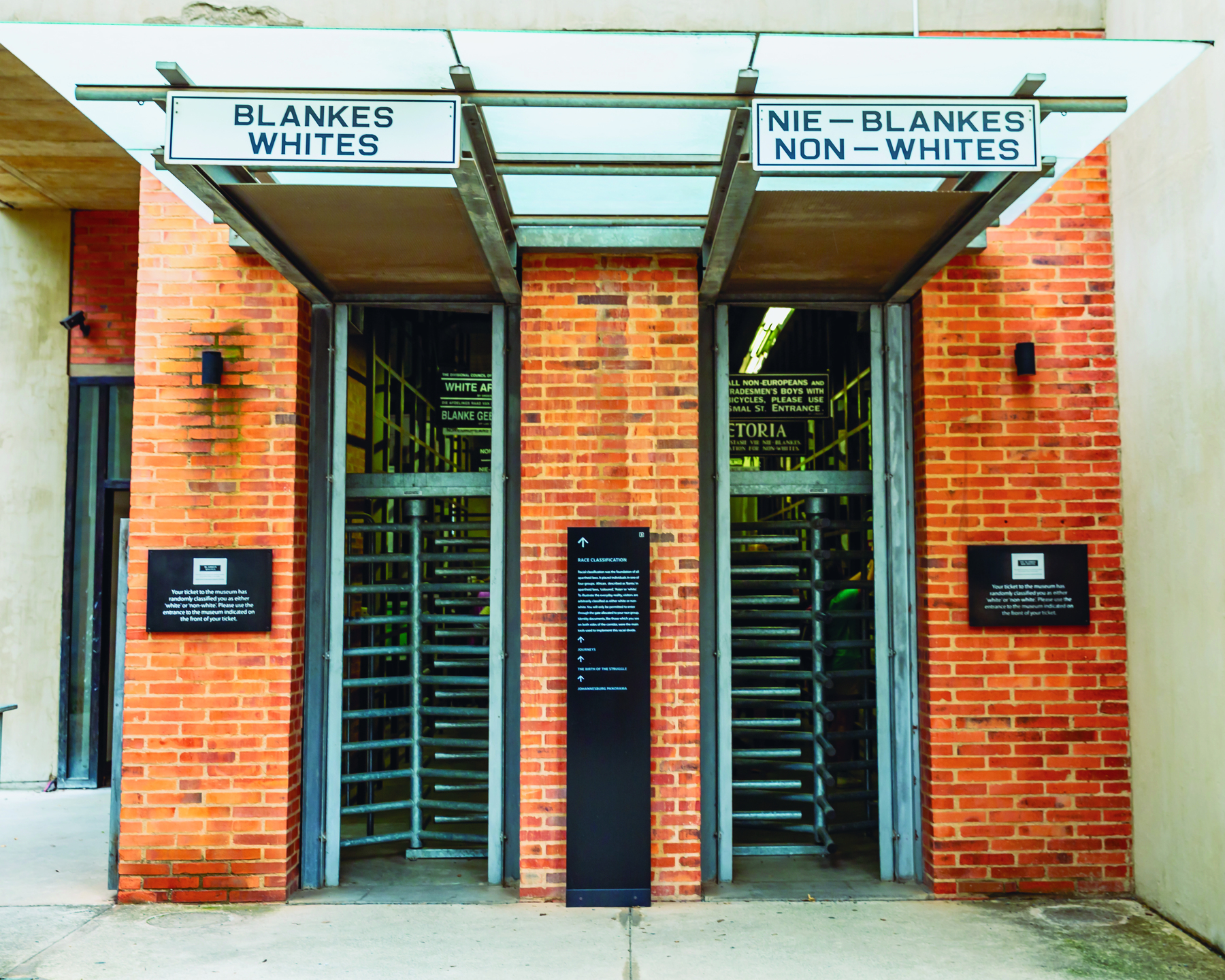 Fotografia. Duas catracas rotatórias com várias barras horizontais em portas de um prédio com uma parede de tijolos. Em uma: blankes, whites. Na outra: nie-blankes, non-whites.