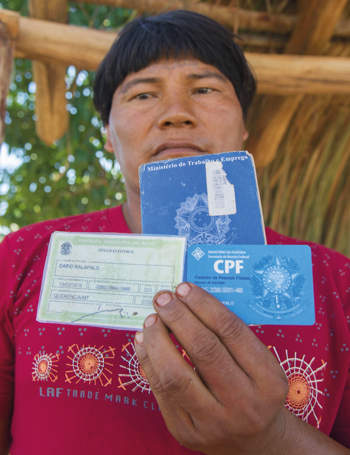 Fotografia. Homem indígena de cabelo preto liso, veste uma camiseta vermelha. Ele segura um documento RG, um CPF e uma carteira de trabalho.