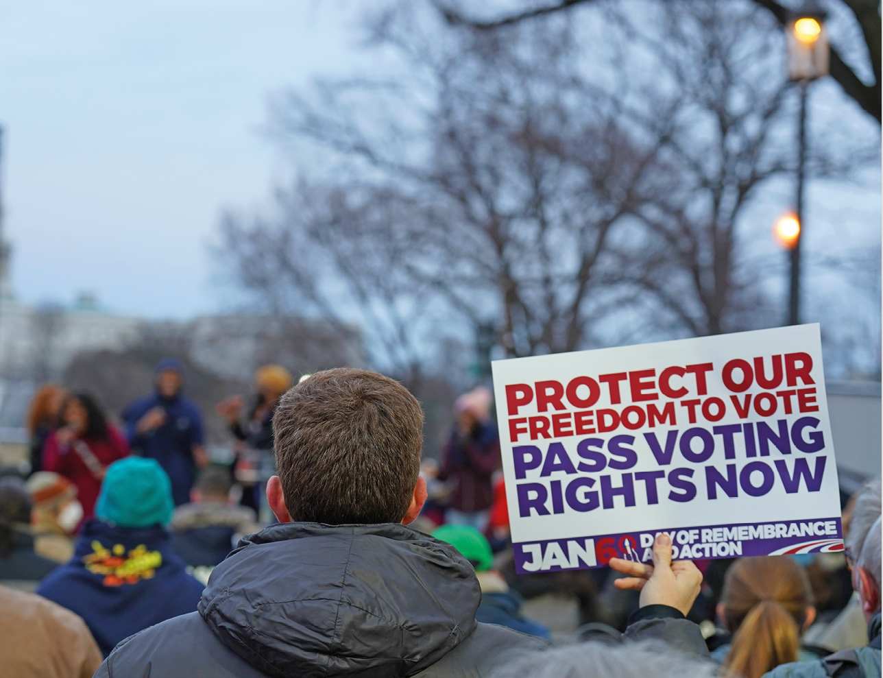 Fotografia. Homem de cabelo castanho liso e jaqueta preta, visto de trás, no meio de outras pessoas em uma manifestação ao ar livre. Ele segura uma placa com o texto: protect our freedom to vote. Pass voting rights now.