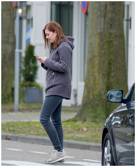 Fotografia. Mulher ruiva de cabelo liso, veste calça e moletom cinza, ela atravessa a rua olhando para o celular que está na sua mão.