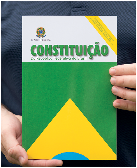 Fotografia. Destaque para uma pessoa segurando um livro. Na capa, parte da bandeira do Brasil e o título: Constituição da República Federativa do Brasil.
