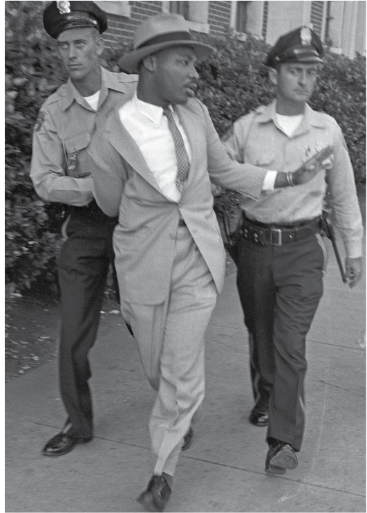 Fotografia. Em preto e branco, homem negro de chapéu, terno e gravata, com o braço direito torcido e preso em suas costas, sendo levado por dois policiais.