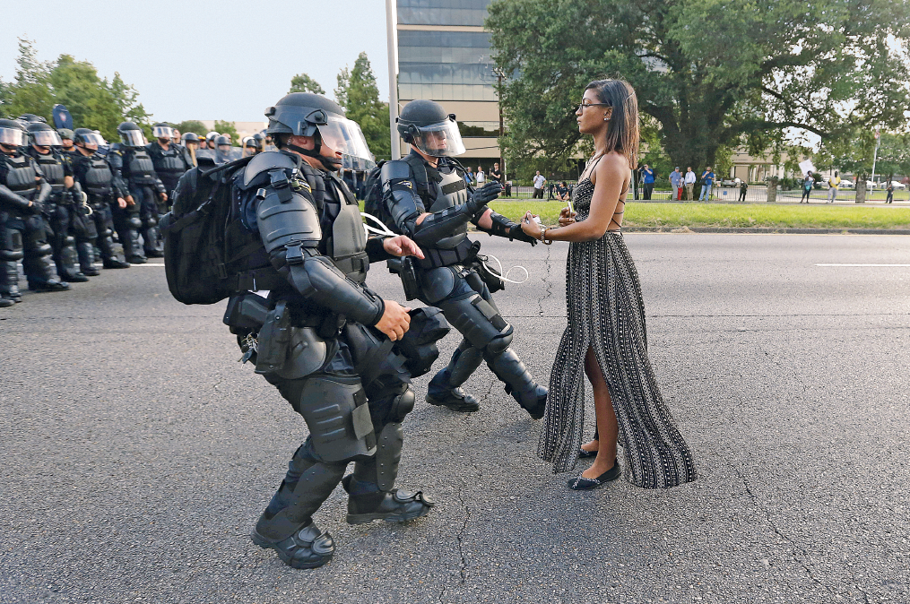 Fotografia. Mulher negra de óculos de grau usando um vestido longo parada em pé na frente de uma tropa de choque que está na rua, com policiais com roupas de proteção, capacetes e armas. Três dos policiais se aproximam dela correndo e agitando os braços.