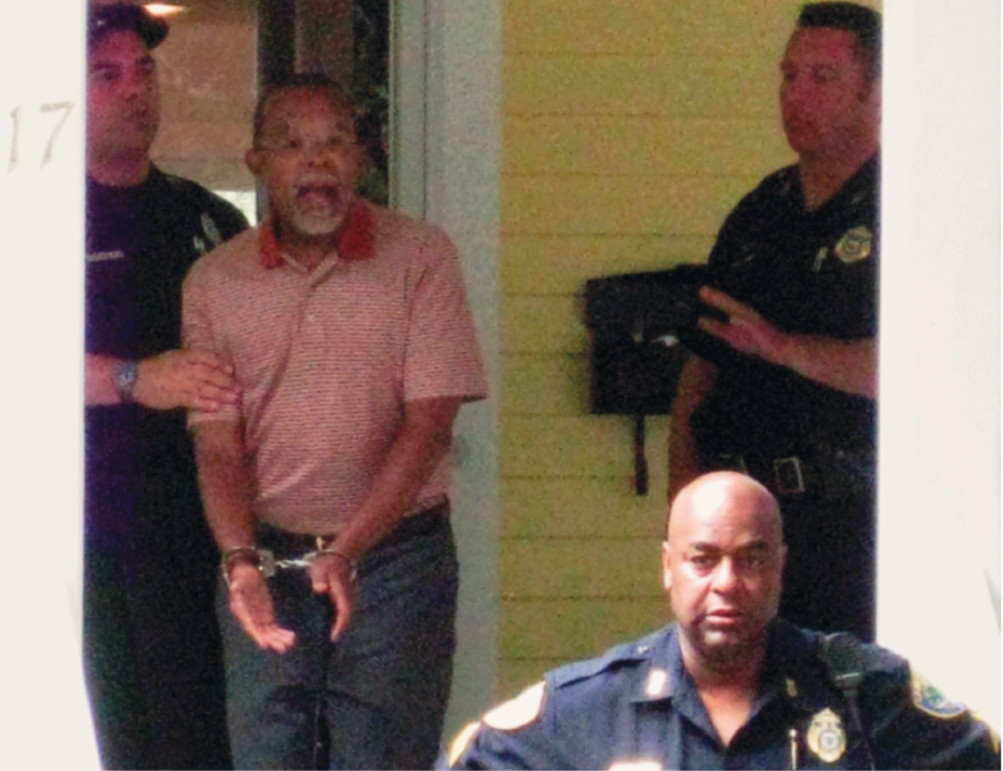 Fotografia. Homem negro de cabelo raspado e camiseta vermelha, com algemas nos pulsos. Ele está sendo segurado por um policial e ao lado há mais dois policiais.