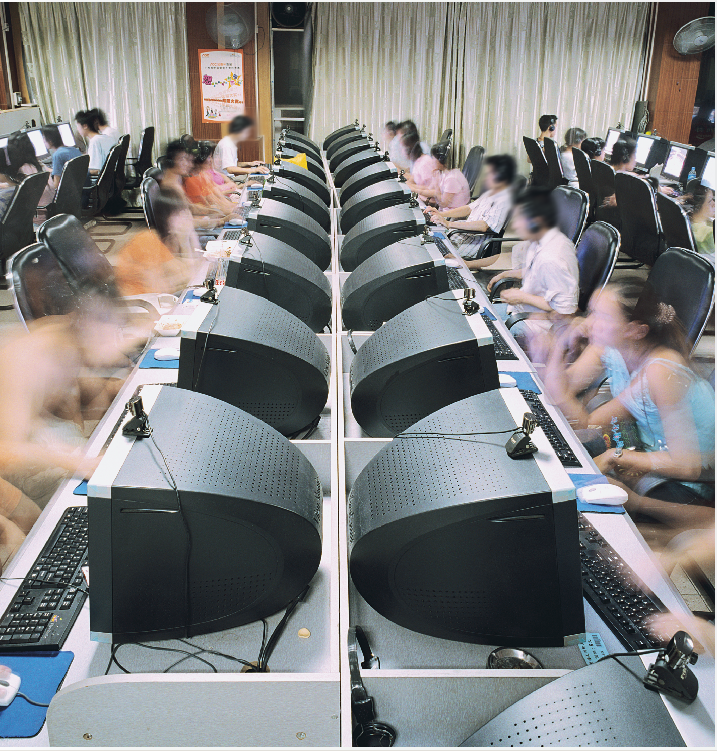 Fotografia. Sala de computadores com dezenas de computadores, e uma pessoa na frente de cada um.