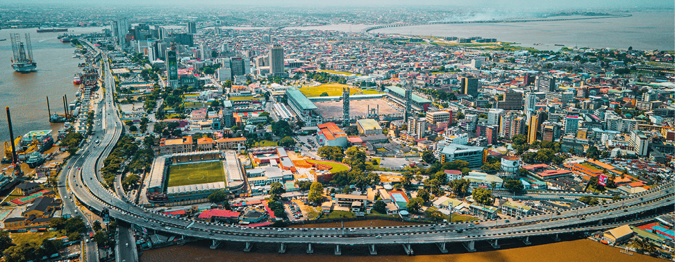 Fotografia. Imagem aérea de uma cidade com vários prédios e um grande viaduto que circula a parte da esquerda indo para a direita.