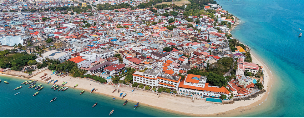 Fotografia. Imagem aérea da ponta de uma cidade litorânea com faixa de areia e mar azul. Há várias casas e construçoes e algumas áreas verdes.