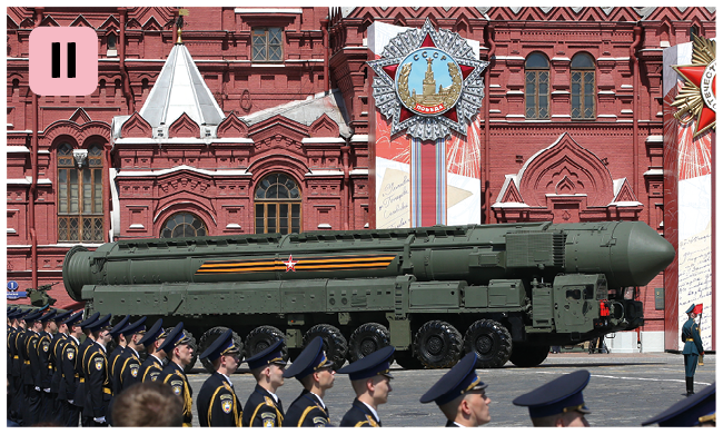 Fotografia. Número dois. Um caminhão verde grande transportando um grande míssil verde na frente de uma construção vermelha e soldados em posição observando.