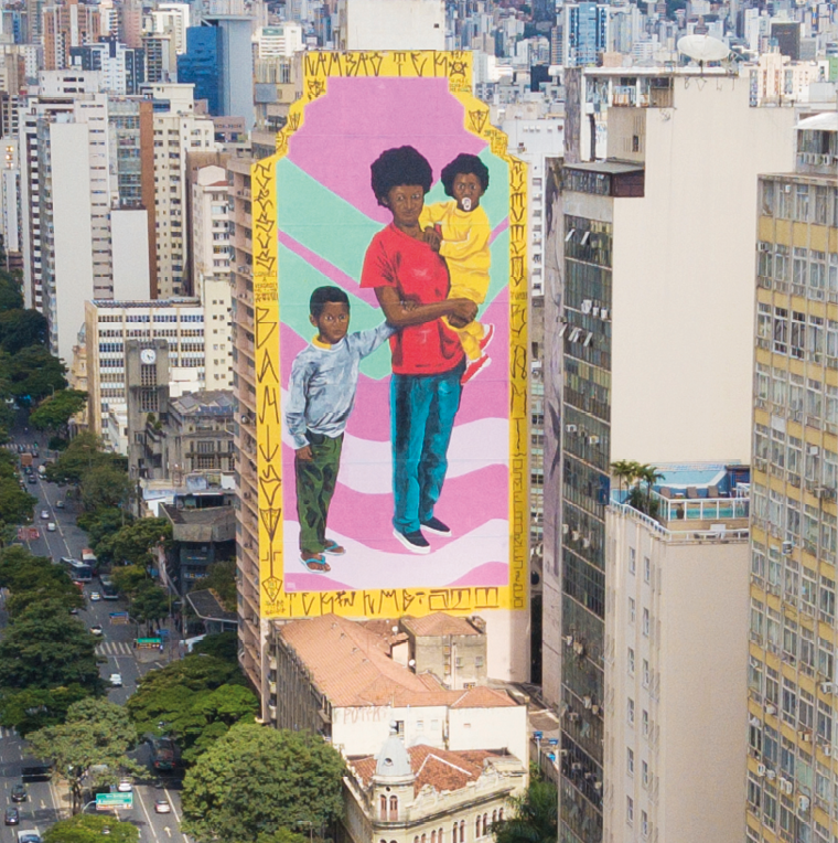 Fotografia. Lateral de um prédio com a pintura de uma mulher negra de camiseta vermelha segurando uma criança de roupa amarela no colo. Ao lado, menino negro  de moletom cinza está segurando a mulher. Fundo na cor rosa com faixas curvas em verde claro e rosa claro.