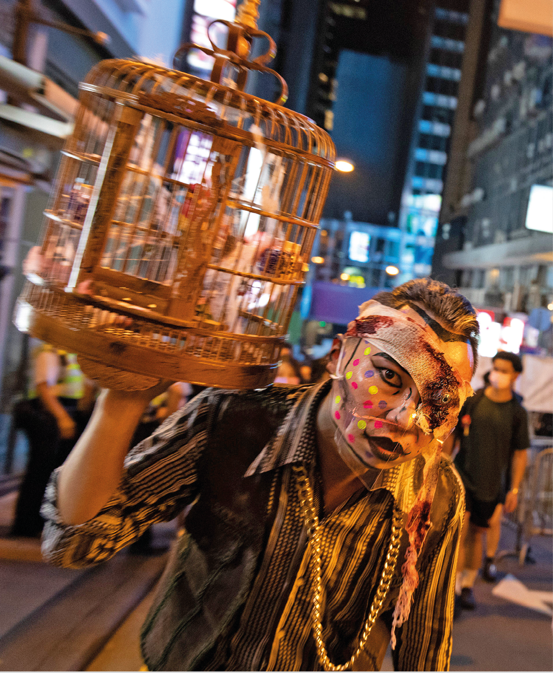 Fotografia. Na rua, à noite, um homem com o rosto pintado e uma máscara. Ele usa uma camisa e uma corrente dourado. Ele segura uma gaiola dourada.
