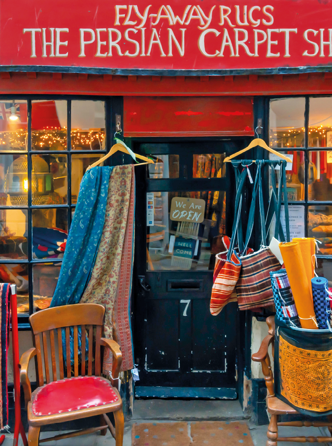 Fotografia. Fachada de uma loja com bolsas e tecidos pendurados em cabides, uma cadeira e uma cadeira com uma cesta com panos coloridos enrolados. Nome da loja: Fly-Away Rugs, the Persian Carpet Shop.