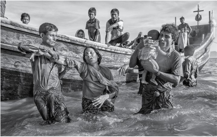 Fotografia. Em preto e branco, homens, mulheres e crianças fora e dentro de um barco. Fora do barco há um homem com uma criança pequena no colo, uma mulher e um rapaz, todos com parte do corpo na água.