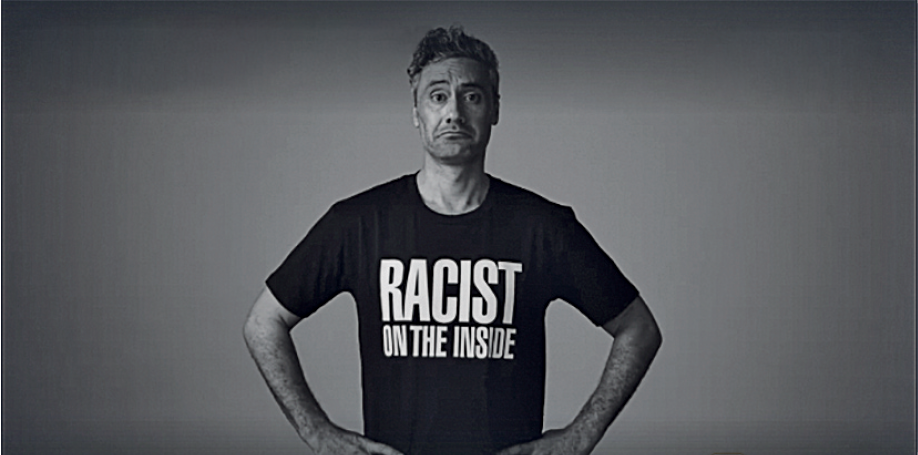 Fotografia. Imagem em preto e branco de um homem com as mãos na cintura. Ele veste uma camiseta preta com o texto: racist on the inside.