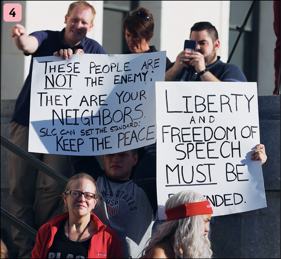 Fotografia. Número quatro. Um grupo de pessoas segurando cartazes. Um deles tem o texto: these people are not the Enemy. They are your neighbors slc can the standarts, keep the peace. O outro diz: liberty and freedom of speech must be, com a última palavra coberta pela cabeça de uma pessoa com cabelos brancos e faixa vermelha na cabeça..