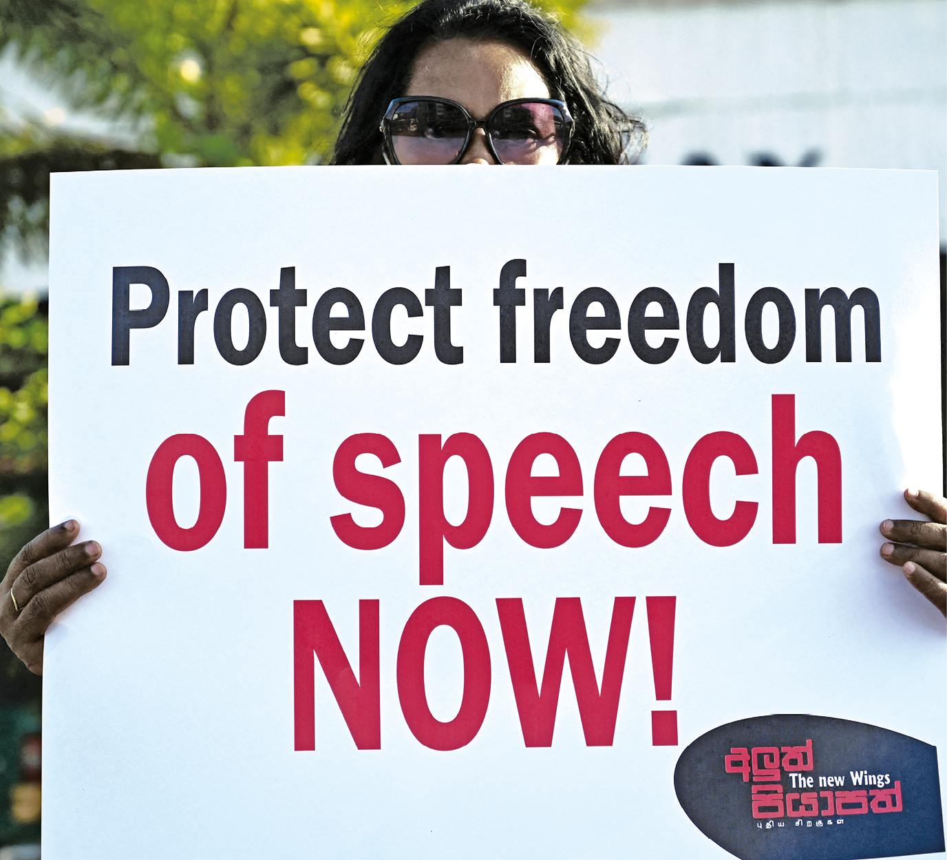 Fotografia. Uma mulher segurando um cartaz branco em frente a seu peito, deixando aparecer apenas os olhos com óculos e as duas mãos, com o texto: protect freedom of speech now!