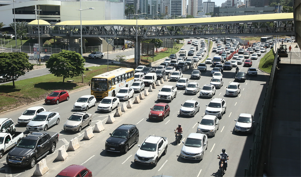 Fotografia. Uma avenida com nove faixas de carros, todos se movendo na mesma direção, com uma barreira de muros pequenos de concreto dividindo a quinta faixa da sexta faixa. Ao fundo, uma passarela coberta com toldo amarelo que atravessa toda a extensão da avenida. Há prédios altos no fundo e algumas árvores ao longo da avenida.