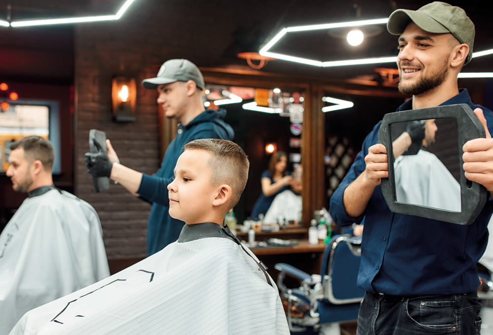 IMAGEM: introdução da unidade 1. um menino está sentado em uma cadeira de salão de cabeleireiro olhando para frente, e atrás dele, o cabeleireiro está segurando um espelho com as duas mãos para mostrar como ficou a parte de trás do cabelo. FIM DA IMAGEM.