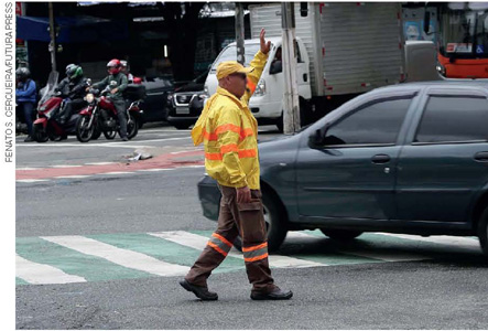 IMAGEM: um homem usando casaco e calças com faixas laranjas fluorescentes está no meio da rua, ao lado de um carro, com o braço erguido e um apito na boca. FIM DA IMAGEM.