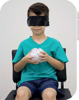 IMAGEM: o menino está sentado com a venda nos olhos, segurando uma bola de papel com as mãos. FIM DA IMAGEM.