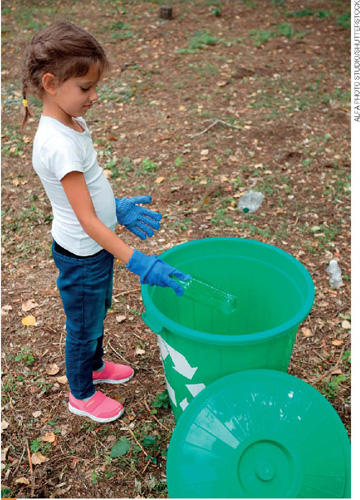 IMAGEM: em um terreno, uma menina usando luvas de proteção coloca uma garrafa de plástico em um cesto de lixo verde. FIM DA IMAGEM.
