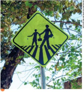 IMAGEM: placa de trânsito com ilustração da silhueta de duas pessoas atravessando na faixa de pedestres. cada uma carrega uma bolsa. FIM DA IMAGEM.