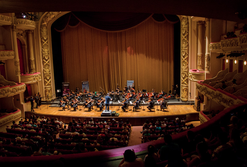 IMAGEM: página dupla. dezenas de pessoas assistem à apresentação de uma orquestra em um teatro com colunas e camarotes enfeitados por arabescos e grandes cortinas ao fundo. FIM DA IMAGEM.