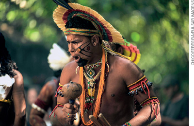 IMAGEM: um homem indígena, trajando adereços tradicionais de sua cultura, toca um chocalho. FIM DA IMAGEM.