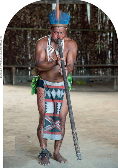 IMAGEM: numa aldeia indígena, um homem da etnia tuyuka toca uma longa flauta que vai da sua boca até o chão. ele usa uma tanga, cocar e outros adereços de sua cultura. FIM DA IMAGEM.