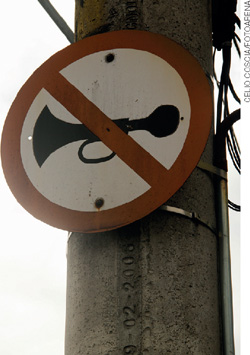 IMAGEM: placa de trânsito com o desenho de uma buzina em seu interior, cortada por uma linha diagonal, indica que é proibido buzinar. FIM DA IMAGEM.