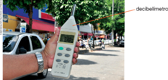 IMAGEM: em uma rua movimentada, um homem segura um decibelímetro. aparelho eletrônico parecido com um controle remoto. FIM DA IMAGEM.