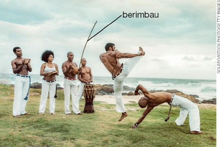 IMAGEM: em uma praia, quatro pessoas tocam instrumentos, sendo um deles o berimbau, enquanto outros dois homens lutam capoeira. todos os seis vestem trajes brancos típicos dessa manifestação cultural. FIM DA IMAGEM.