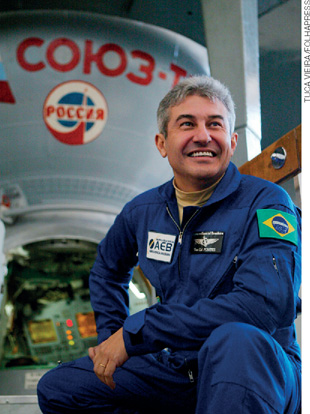 IMAGEM: retratado do astronauta brasileiro, marcos pontes, sorridente em frente a uma sonda espacial. ele veste um macacão espacial com a bandeira do brasil pregada no ombro. FIM DA IMAGEM.