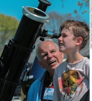 IMAGEM: em um ambiente aberto, um menino espia um telescópio portátil, sobre a supervisão de um homem idoso. FIM DA IMAGEM.